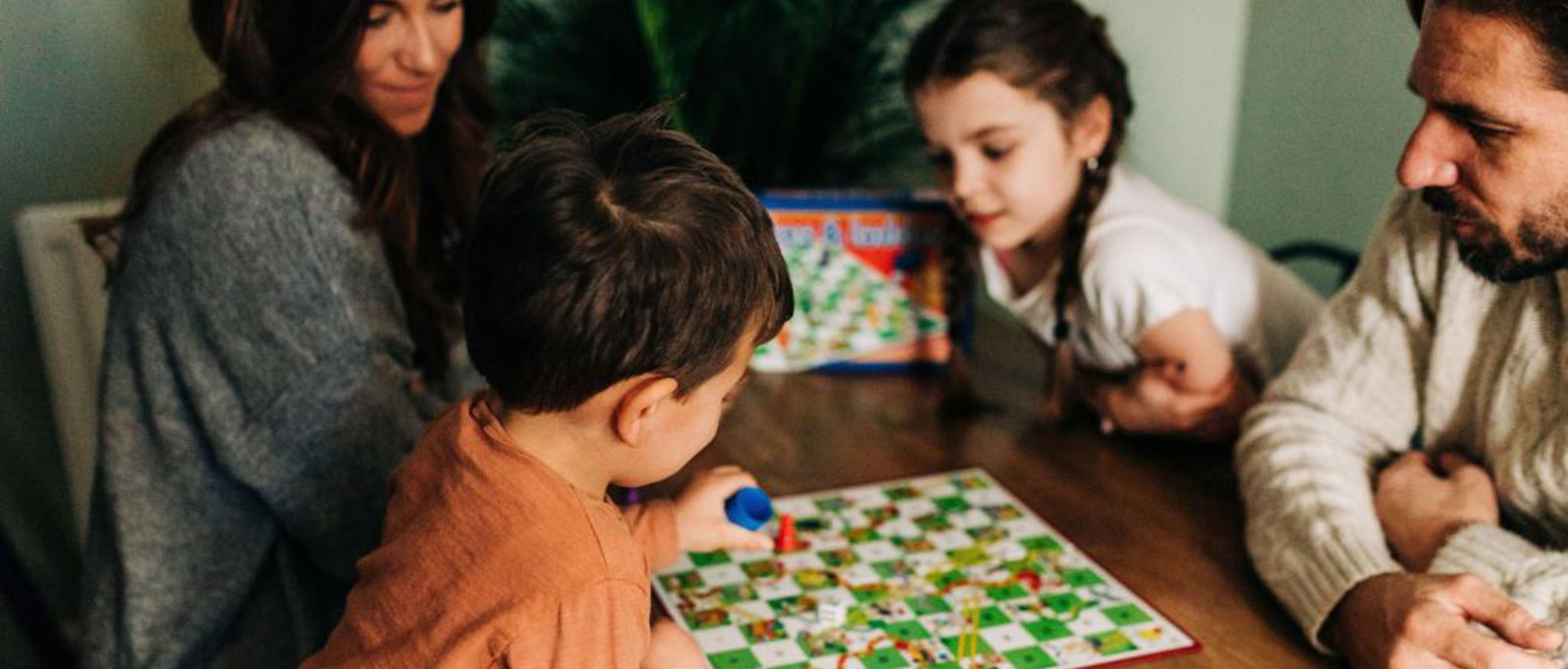 A family is sitting around a table playing M.Y Snakes and Ladders together