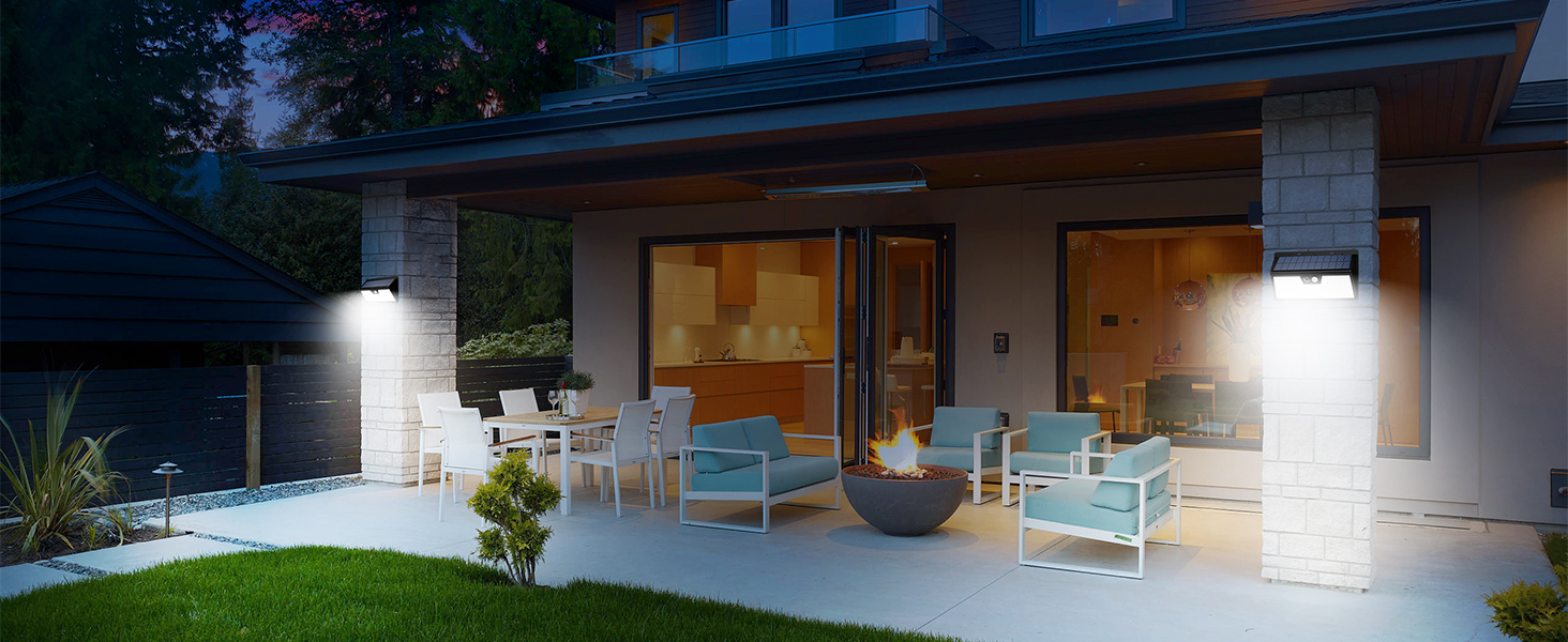 Outdoor patio area with modern furniture, including chairs and a fire pit. White exterior walls with large glass doors. Illuminated by wall-mounted lights and warm interior glow.