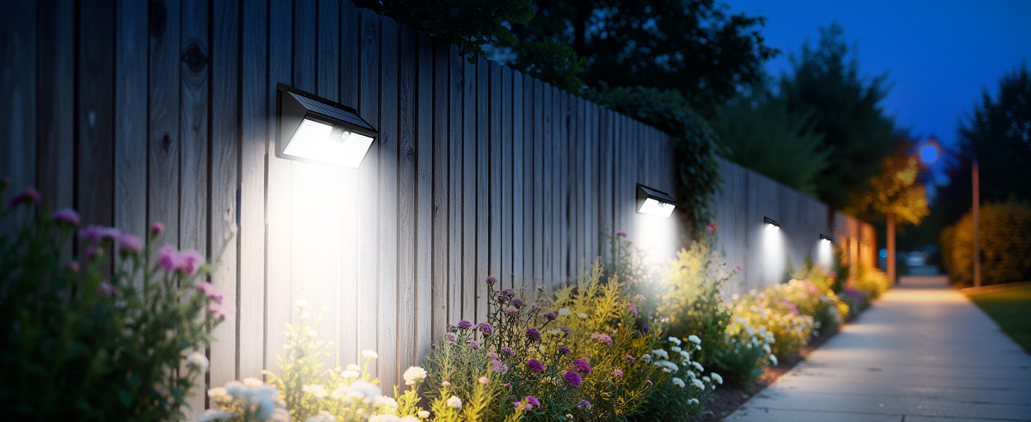 Row of outdoor wall-mounted lights illuminating a garden pathway at dusk, with flowering plants visible along wooden fence panels.