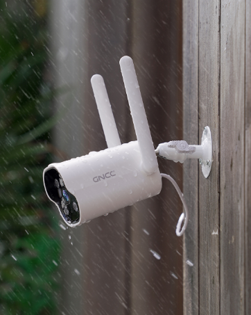 Small white circular indoor security camera on wooden surface near window, with large pinecones in foreground. Camera features a dark lens and appears to be plugged in.