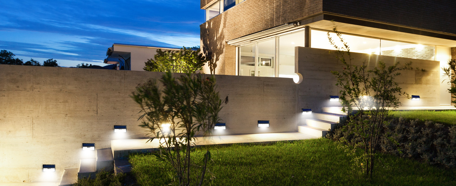 Outdoor LED wall lights illuminating a modern house exterior at night. Lights are mounted along a concrete wall, casting a warm glow on the landscaping and steps.