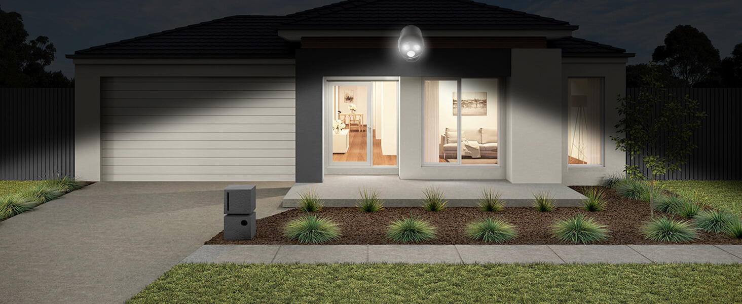 Modern single-story house at night with illuminated exterior. White walls, large garage door, manicured front garden, and bright porch light visible.