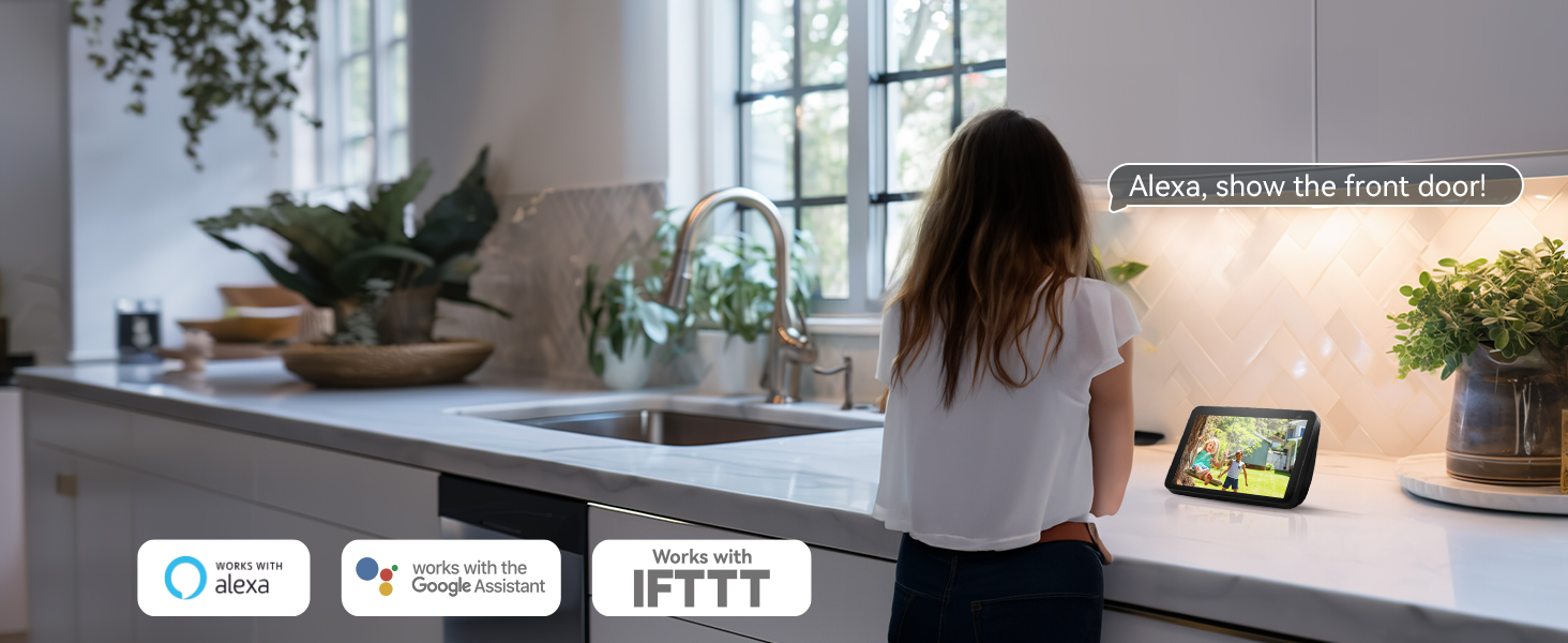 Smart home device on kitchen counter. Screen displays message, icons show compatibility with Alexa, Google Assistant, and IFTTT. Woman interacting with device in background.