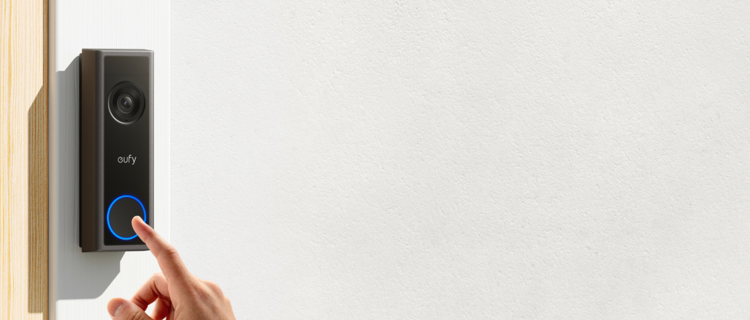 Doorbell camera with person's finger pressing illuminated blue ring button. Black rectangular device mounted on light-colored wall beside wooden door frame.