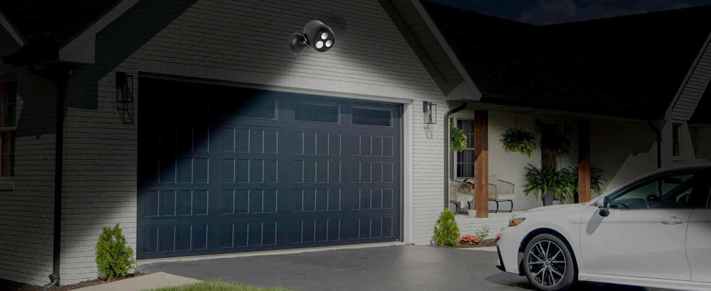 Nighttime view of a house exterior with large blue garage door. White trim and siding visible. Car parked in driveway, plants near entrance.
