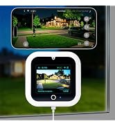 Smartphone displaying outdoor nighttime landscape with illuminated house and green lawn, positioned above two white cylindrical smart home devices with power cords.