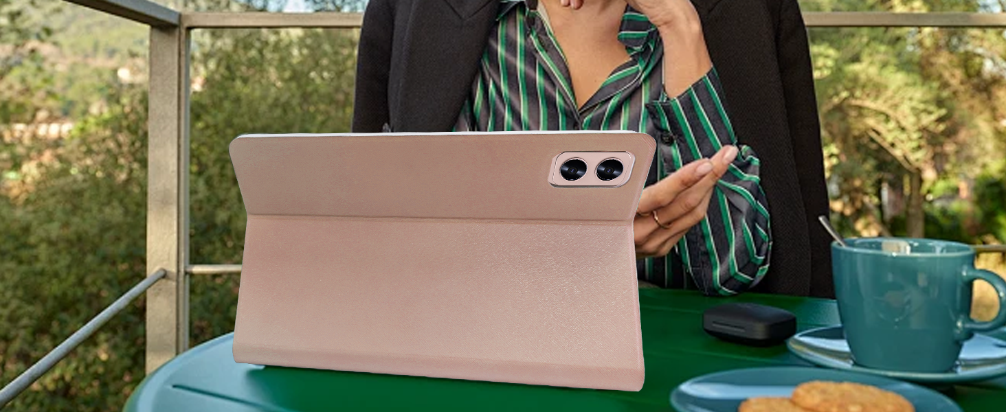 Rose gold tablet with dual rear cameras, shown in use outdoors. Person holds device in landscape orientation on a green table with a teal mug visible.