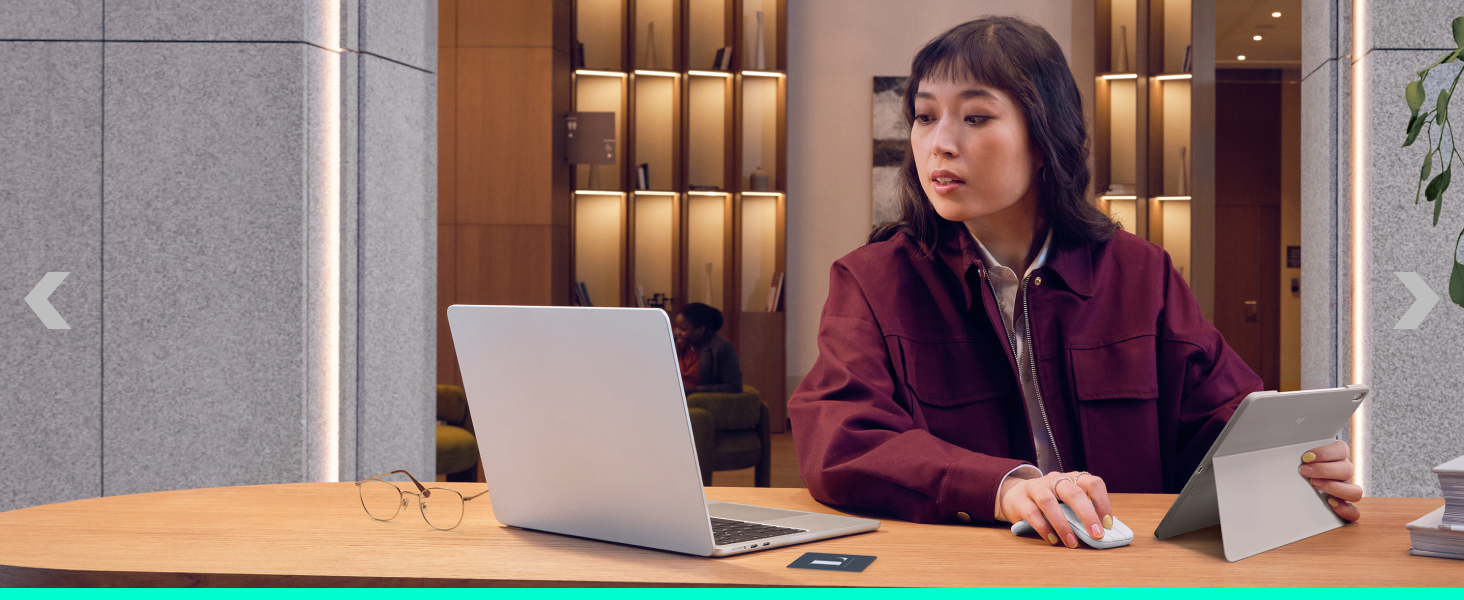 Woman in burgundy cardigan working at laptop in bright office setting with shelving and bookshelves visible in background.
