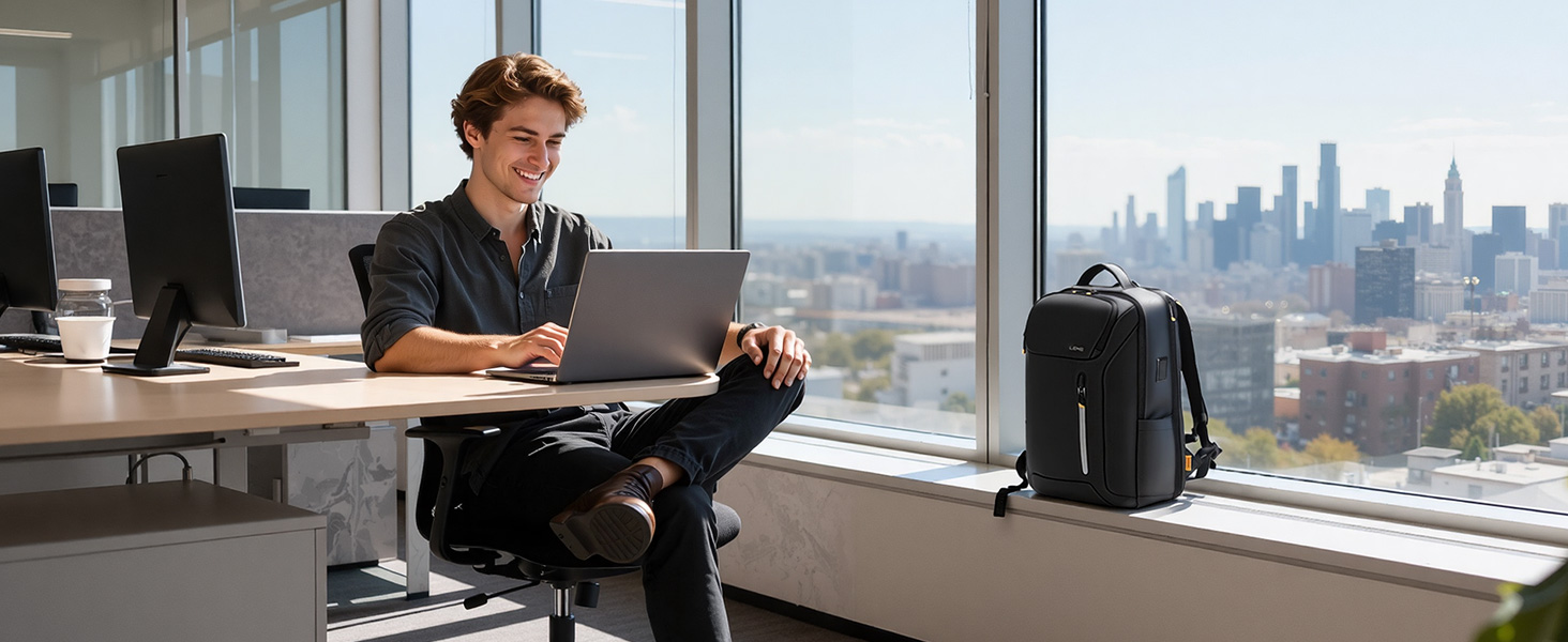 Man in dark business casual attire sitting at desk using laptop with cityscape visible through large windows behind him. Black backpack placed on floor beside desk in modern office environment.