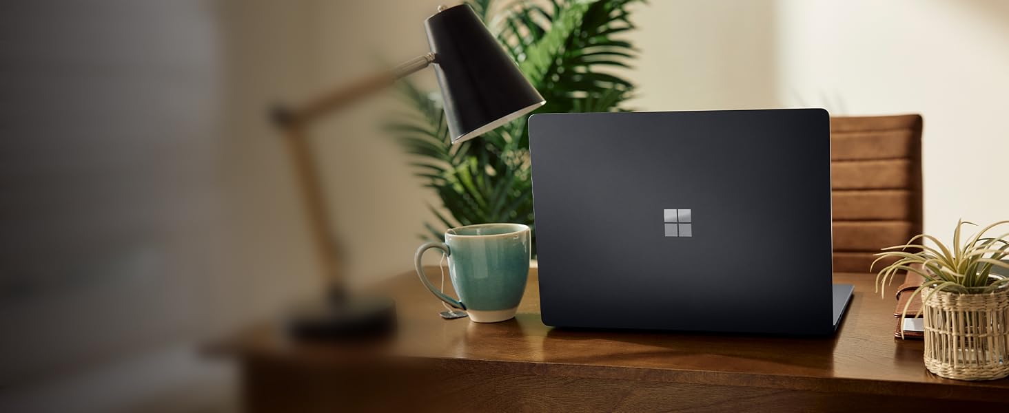 Black Microsoft Surface laptop positioned on wooden desk with potted green plant and blurred background, showcasing device in home office setting.