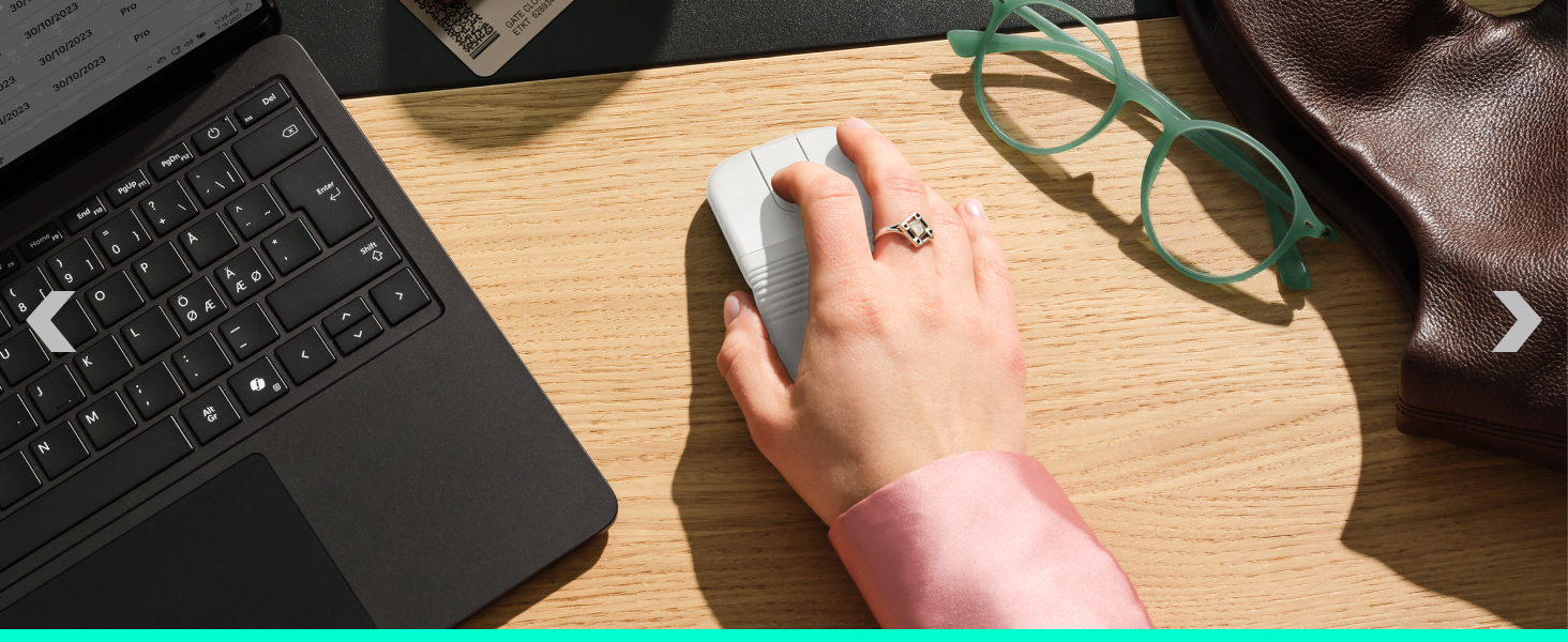 Hand wearing a ring rests on wooden desk surface next to laptop keyboard, eyeglasses, and leather item in overhead flat-lay composition.