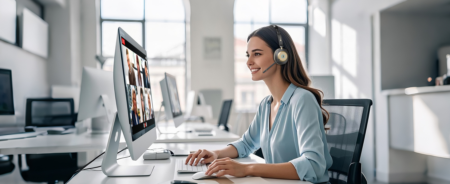 Modern office workspace with iMac computer display showing video conference. Person wearing white headset seated at desk in bright, open-plan office environment.