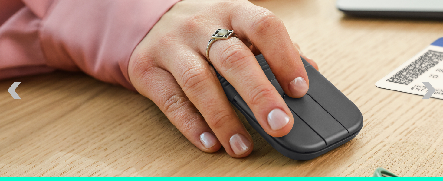 Hand using gray wireless computer mouse on wooden desktop surface with papers and keyboard visible in background.