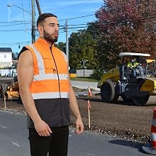 High-visibility safety vest worn by a person at a road construction site. Yellow vest with reflective strips, black lower half, and full front zipper.
