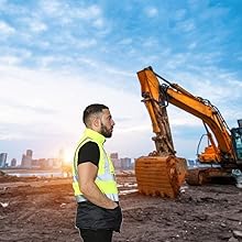 Construction worker in high-visibility vest standing near road paving equipment. Background shows asphalt laying machine and steamroller on a newly paved road.