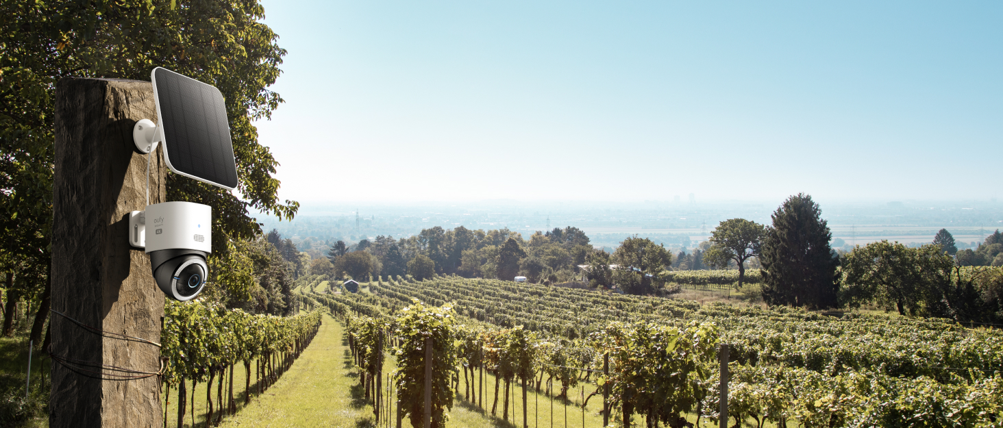 Outdoor security camera mounted on a wooden post overlooking a vineyard. Rolling hills and trees visible in the background under a blue sky.