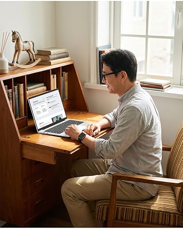 Woman at desk wearing glasses and dark blazer, using pink laptop and wireless mouse on white workspace with notebook and pen visible.