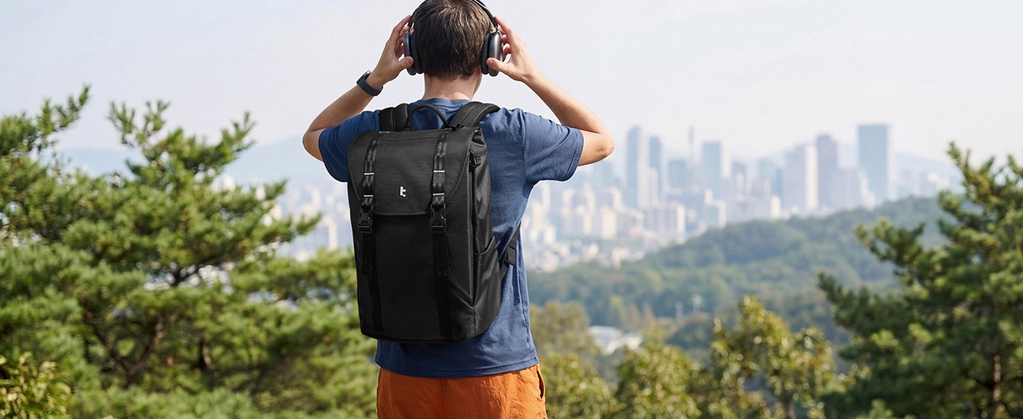 Person wearing black backpack viewed from behind, standing outdoors overlooking city skyline. Green vegetation in foreground, urban landscape in distance.