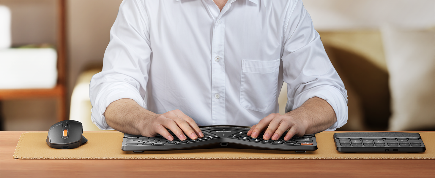 Man in light blue shirt seated at wooden desk, hands positioned on black computer keyboard with mouse and small device on desktop workspace.