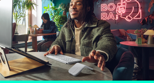Person wearing olive-green jacket at desk in modern workspace with neon pink sign reading 'BOTAI' visible in background. Indoor scene showing professional setup with laptop and desk.