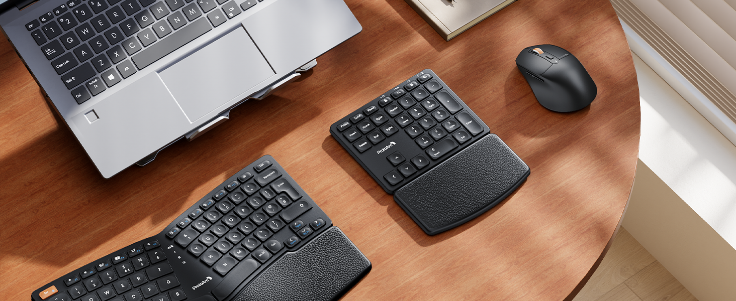 Ergonomic split keyboard on wooden desk, alongside laptop, wireless mouse, and separate number pad. Keyboard features curved design and wrist rests for comfort.