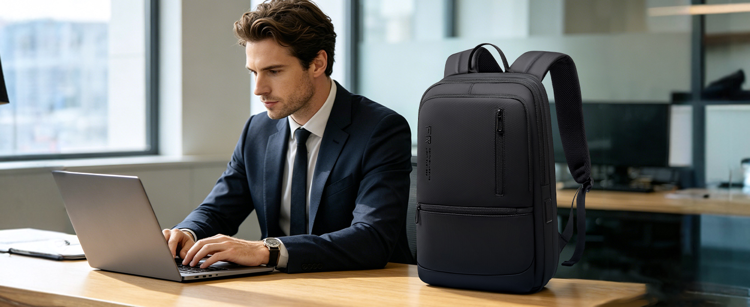 Man in business suit and tie working at desk with laptop. Black backpack positioned beside him. Product demonstrated in professional office workspace setting.