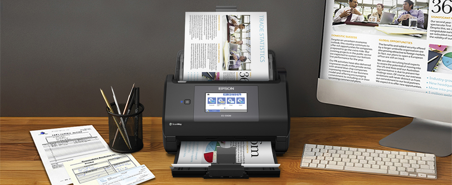 Compact black desktop scanner positioned on desk between papers and office supplies, with monitor and keyboard visible in organized workspace setup.