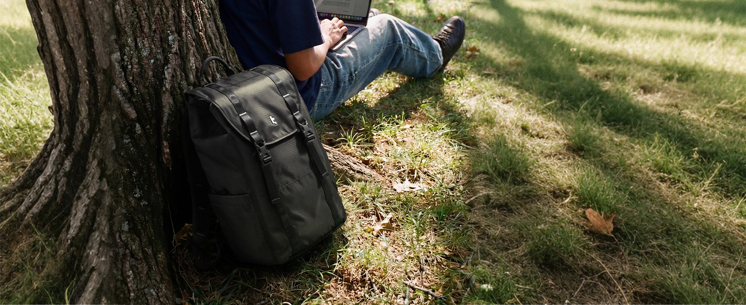 Person wearing dark drawstring shorts, seated outdoors against tree on grass with backpack nearby.