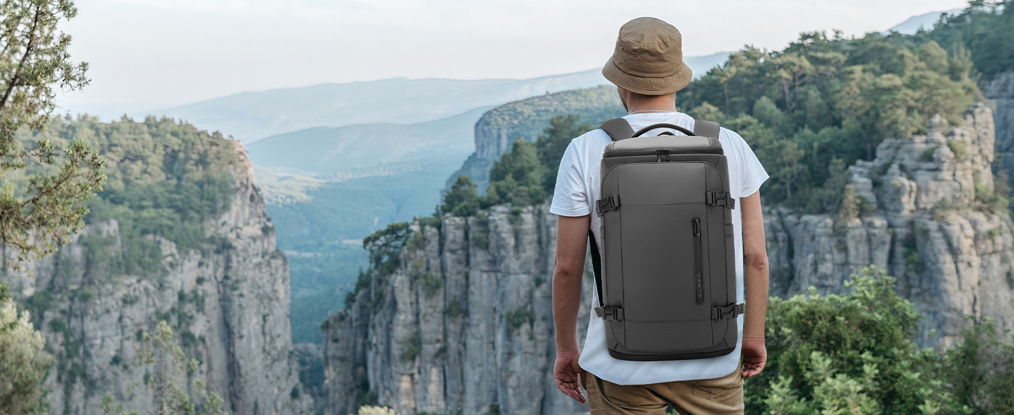 Person wearing gray backpack with white shirt standing on rocky mountain overlook with layered cliff formations and forested valley in background during daytime.