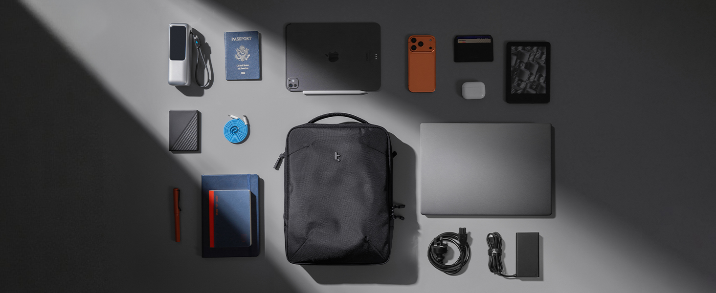 Flat lay arrangement of travel essentials including black backpack, wallet, passport, phone, chargers, keys, sunglasses, and various small accessories on dark background.