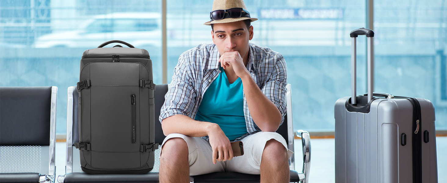 Man in blue apron sitting on luggage against airport backdrop with gray soft-sided carry-on backpack and hard-case rolling suitcase. Travel luggage and bag collection display.