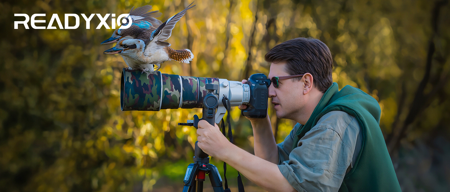Text reads 'READYXIO'. Photographer using professional camera with telephoto lens mounted on tripod, photographing outdoors with blurred autumn foliage in background.