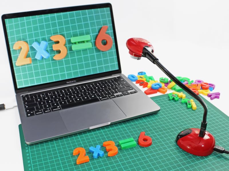 Laptop displaying math equation on green grid background. Colorful plastic numbers and symbols scattered nearby. Red desk lamp in foreground.