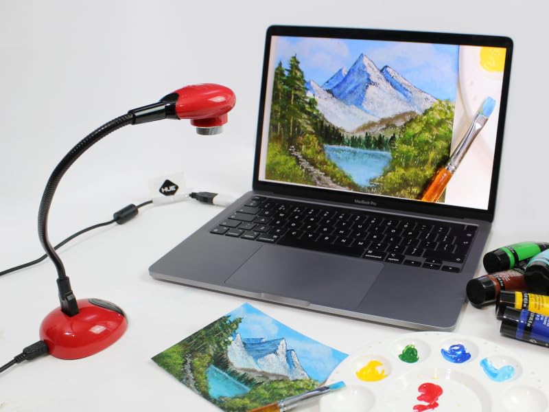 Desk setup with laptop displaying mountain landscape, red desk lamp, painting supplies including palette with colorful paints, and brushes for digital art creation.