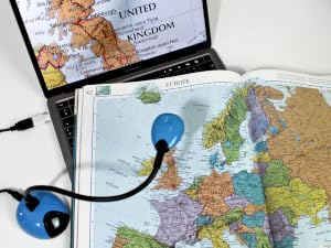 Open laptop displaying map of United Kingdom, alongside paper map of Europe. Blue mouse-shaped device rests on the paper map.