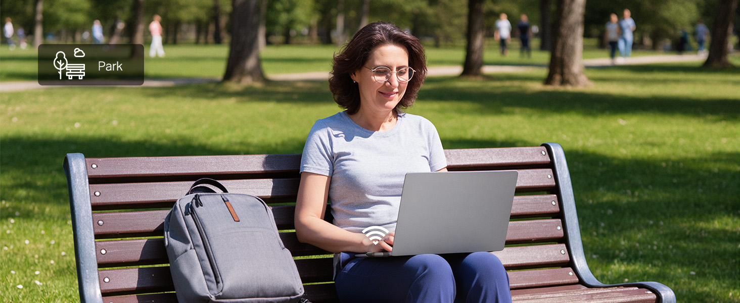Woman in white t-shirt using laptop while sitting on outdoor park bench, with gray shoulder bag beside her and green grass and trees in background.