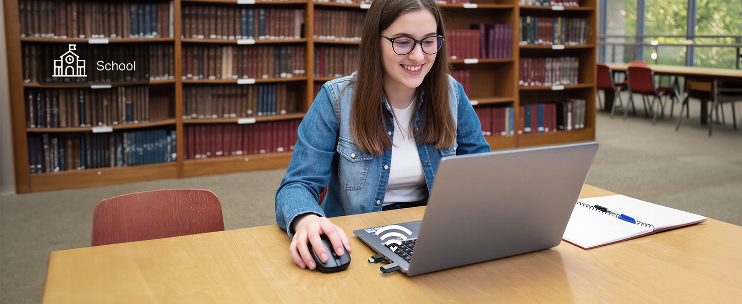Woman wearing glasses and blue denim shirt using laptop at wooden table in library with bookshelves visible behind her.