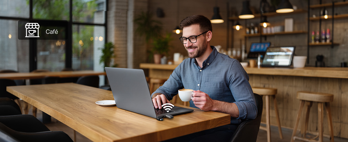 Man wearing glasses and blue shirt seated at wooden table, holding and viewing laptop. Indoor workspace setting with blurred background.